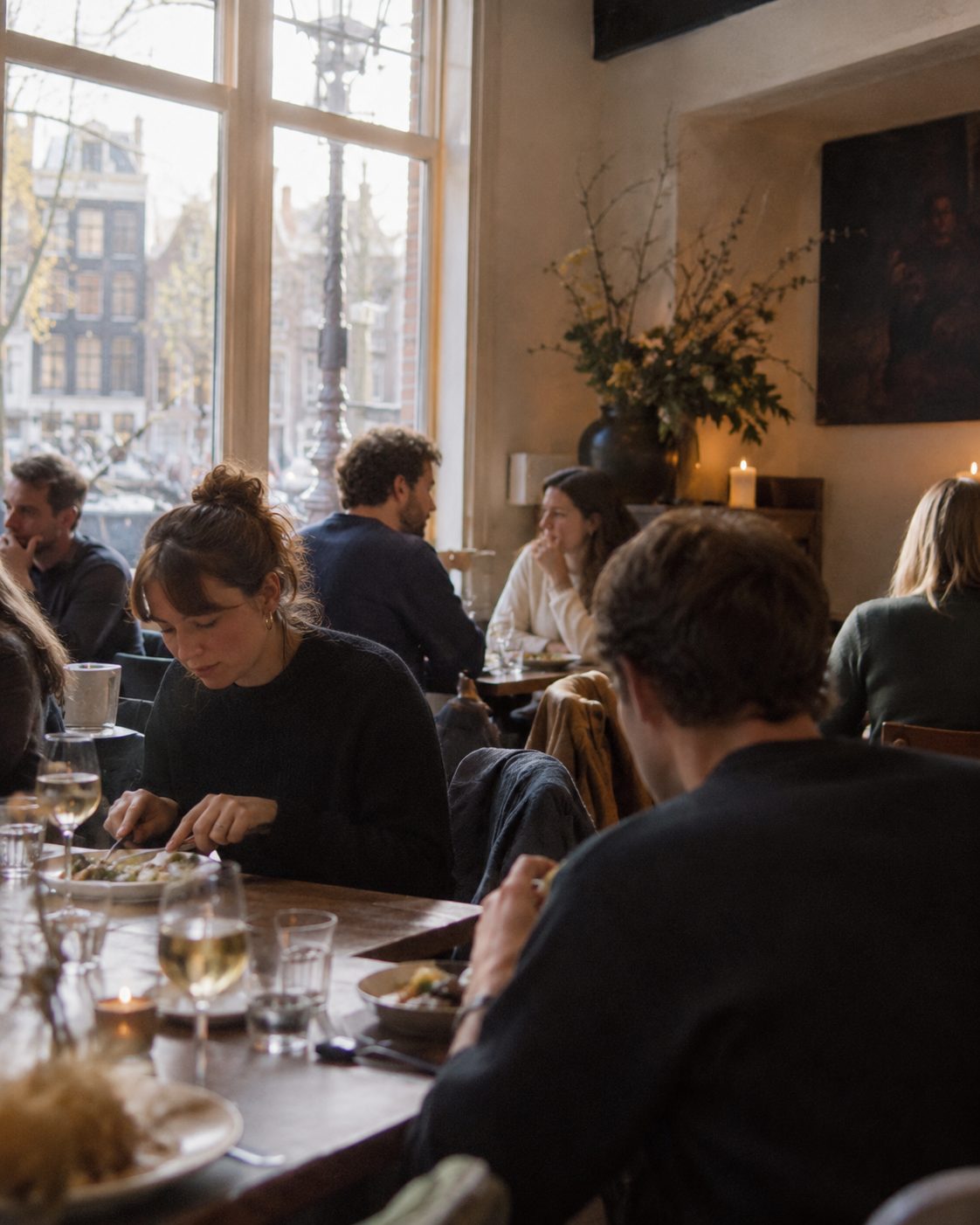 Gasten aan tafel in een vol restaurant, warm licht door de ramen.